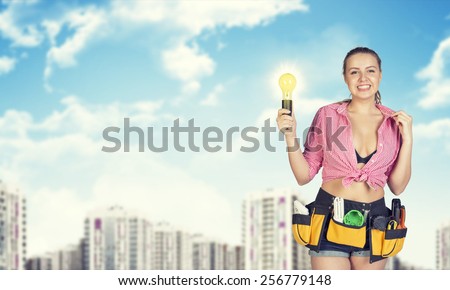 Woman in tool belt with different tools holding light bulb. Blurred buildings in background