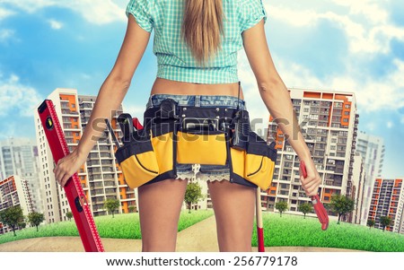 Woman in tool belt with different tools stands back, holding building level and wrench. Cropped image. Green hill with road and buildings on background