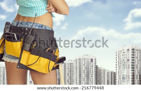 Woman in tool belt with different tools stands back. Cropped image. Buildings and sky as backdrop