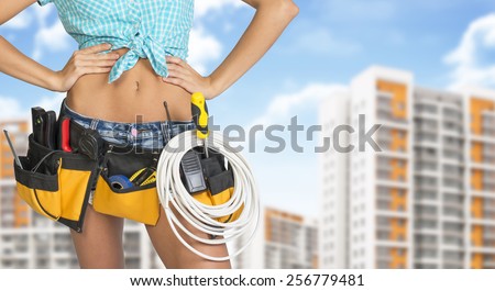 Woman in tool belt with different tools. Hands on hip. Cropped image. Buildings and sky on background