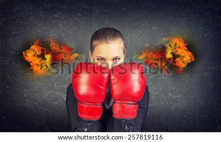 Woman in suit and boxing gloves covers her face. Fire from ears. Concrete gray wall as backdrop