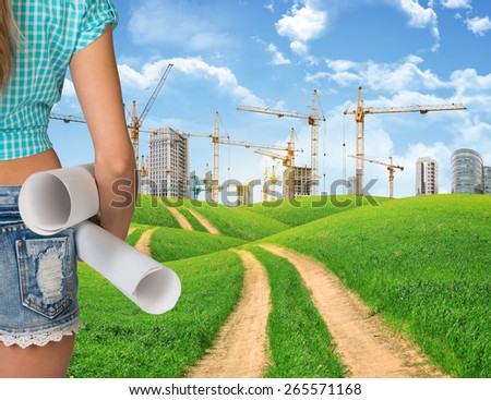 Girl builder in jeans shorts holding paper scrolls, cropped image. Green hills, country road and buildings with tower cranes as backdrop