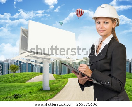 Businesswoman in suit and helmet holding clipboard, looking at camera. Large billboard, road, grass hills and city as backdrop