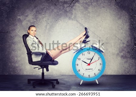 Businesswoman sits in chair, looking at camera. Put your feet up on big red alarm clock. Concrete background