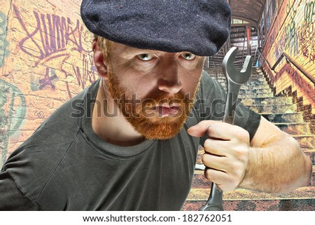 Angry villain plumber in hat with red beard  threatening with a wrench in composite background of underground passage covered with graffiti