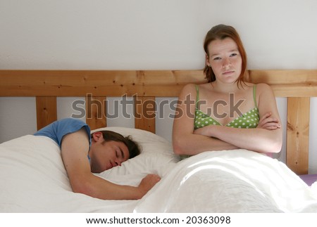 A couple in bed, the man is sleeping lazily, the woman looks disappointed about him.