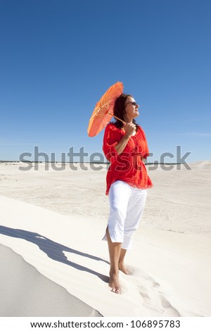 An attractive and confident looking mature woman with red blouse and umbrella, isolated on white sand dune overlooking a desert panorama, with clear blue summer sky as background and copy space.