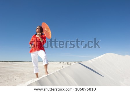 An attractive and confident looking mature woman with red blouse and umbrella, isolated on white sand dune overlooking a desert panorama, with clear blue summer sky as background and copy space.