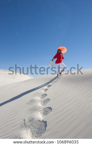 Upward view of attractive and happy looking mature woman walking up a white sand dune, leaving footsteps, with red blouse, matching umbrella, isolated with clear blue sky as background and copy space.
