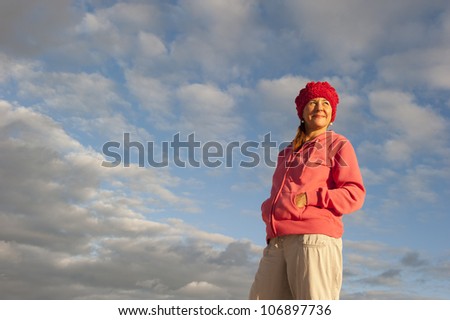 Portrait of a beautiful looking middle aged woman in casual pose, wearing pink sweater and red cap, with soft sunlight on her face and picturesque cloudscape as background and copy space.