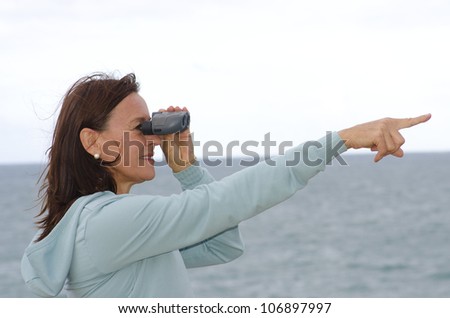 A pretty looking middle aged woman with binoculars standing at seaside and pointing with one arm towards the distance to the right, with ocean and bright sky as blurred background and copy space.