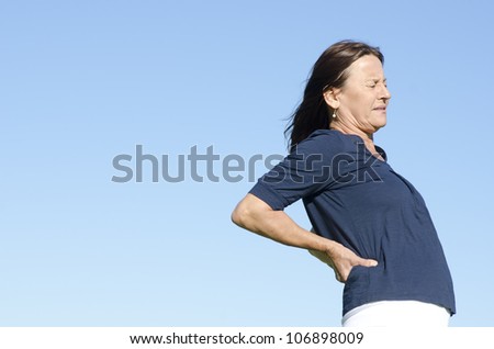 A mature woman in pain, obviously suffering from back problems, with a stressful facial expression, clear blue sky as background and copy space.