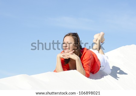 An attractive looking middle aged woman, wearing a bright red blouse, is lying relaxed and happy on top of a white sand dune, with blue summer sky as background and copy space.