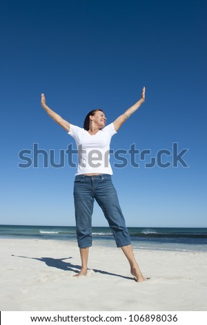 An attractive looking mature woman in her fifties is enjoying early retirement with a day on the beach, standing with her arms up, wide open ocean and clean blue sky as background and copy space.