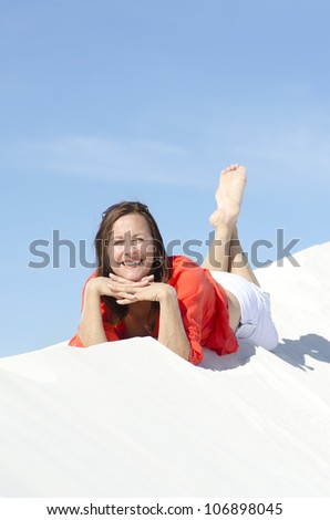 A pretty looking middle aged woman, wearing a bright red blouse, is lying relaxed and happy on top of a white sand dune, with blue summer sky as background and copy space.