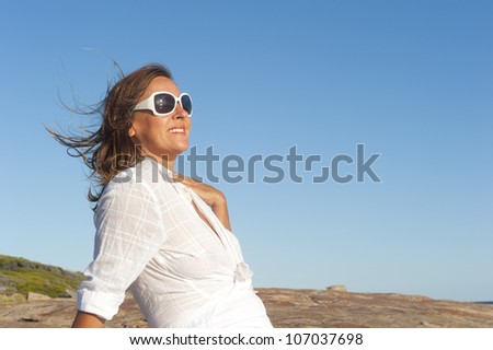 Portrait of an attractive middle aged woman, smiling and relaxed, wearing white blouse and sunglasses, isolated with blue sky as background and copy space.