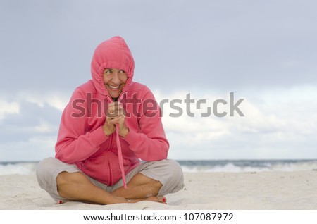 Portrait of happy relaxed and pretty looking mature woman with hooded pink sweater, keeping warm at beach, isolated with dark storm clouds and ocean as background and copy space.