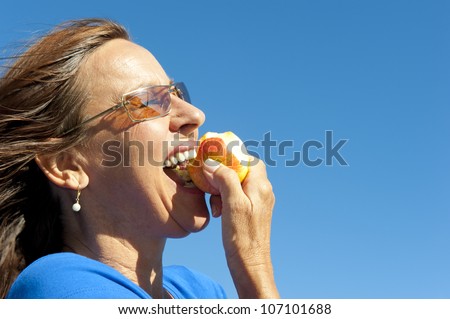 Portrait of happy woman keeping healthy and fit by eating fresh fruits, biting in apple, isolated with blue sky as background and copy space.