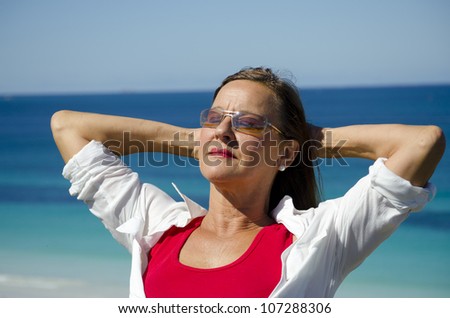 Portrait of an attractive looking mature woman with arms relaxed behind head, isolated with ocean and blue sky as blurred background and copy space.
