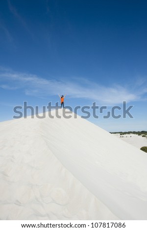 An attractive girl is standing alone on a white sand dune in a winning pose with her arms high up towards the clear blue sky, surrounded by scenic desert landscape, lots of copy space.