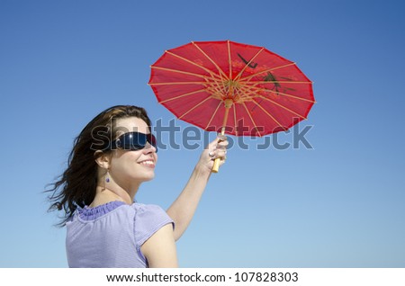 A pretty looking young woman is posing on a sunny day with a red umbrella in one hand, with clear blue summer sky in the background and lots of copy space.