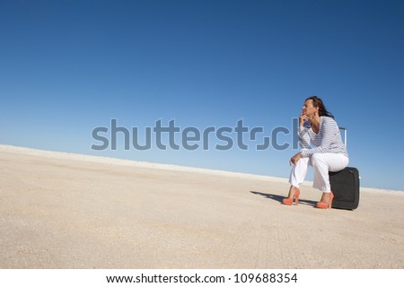 Sexy looking mature woman sitting lost in a remote desert area with a suitcase, looking for a transport to travel, isolated with blue sky as background and copy space..