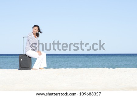 Attractive lady on holiday sitting on suitcase alone at a beach overlooking the sea, isolated with ocean and blue sky as background and copy space.