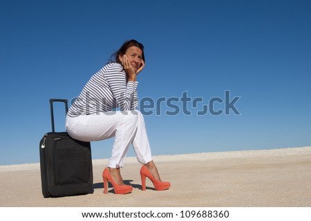 Sexy looking mature woman sitting lost in a remote desert area on a suitcase, looking for a transport to travel, isolated with blue sky as background and copy space..