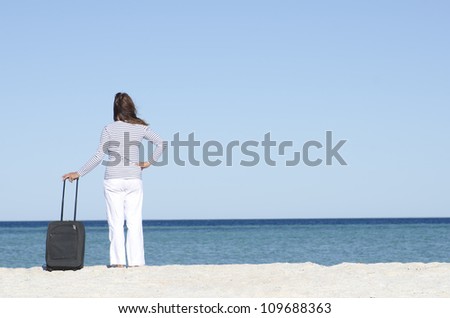 Attractive lady on holiday standing with suitcase alone at a beach overlooking the sea, isolated with ocean and blue sky as background and copy space.