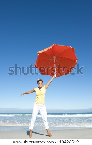 A pretty looking mature woman dancing happy and cheerful with big orange umbrella at beach, isolated with ocean and blue sky as background and copy space.
