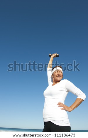 Attractive and active senior woman exercising with weights at the beach, with ocean and blue sky as background and copy space.