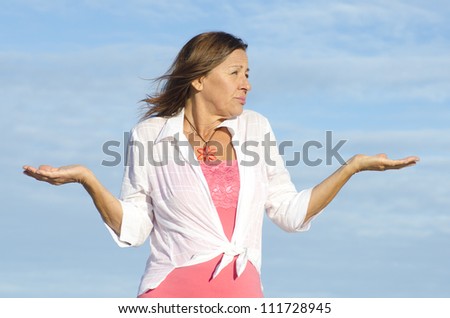 Mature woman shrugging innocent and confused shoulders, gesturing with arms up, isolated with blue sky as background and copy space.