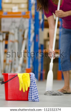 Business cleaning of industrial warehouse with isolated red bucket, yellow glove and legs of female cleaner in blurred background and copy space.