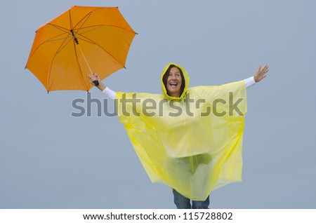 Portrait mature woman standing in the rain, wearing yellow raincoat and orange umbrella, isolated with grey sky as background and copy space.