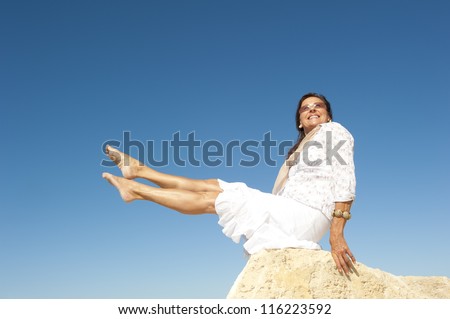 Attractive and fit and healthy looking middle aged woman sitting on rock with legs up in air, isolated with blue sky as background and copy space.
