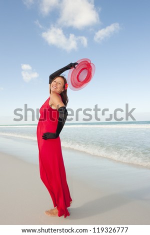 Beautiful looking lady posing happy smiling in elegant red dress and long black gloves at beach, isolated with ocean and blue sky as background and copy space.