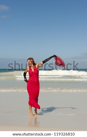 Sexy looking lady posing happy smiling in elegant red dress and long black gloves dancing at beach, isolated with ocean and blue sky as background and copy space.