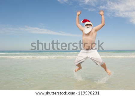 Happy joyful and cheerful Santa Claus on summer holiday, jumping in shallow water at tropical beach, isolated with ocean and blue sky as background and copy space.
