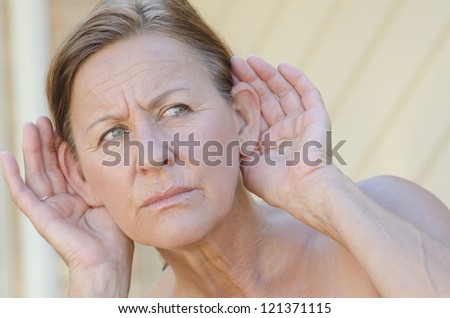 Portrait beautiful looking mature woman looking worried while listening with hands close to ears, isolated outdoor with blurred background.