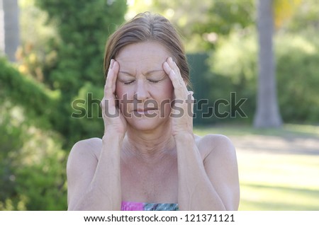 Outdoor portrait of concerned mature woman with headache and hands at forehead, isolated with blurred background.