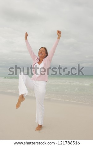 Portrait Happy beautiful middle aged woman smiling joyful and cheerful arms up at beach holiday and active retirement, isolated with ocean and overcast sky as blurred background and copy space.