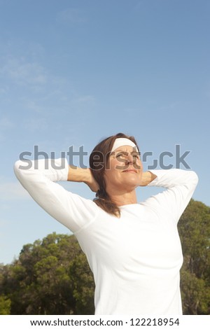 Active and fit attractive middle aged woman in white shirt and headband exercising happy in park with arms up, isolated with trees and blue sky as background and copy space.