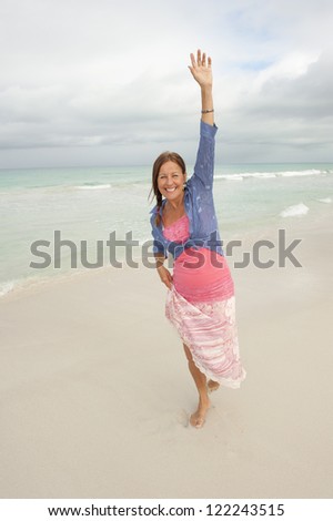 Beautiful middle aged woman with arms up happy and confident at beach vacation enjoying active retirement, isolated with ocean and cloudy sky as background and copy space.