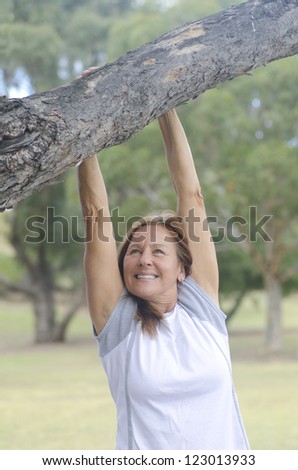 Attractive and happy looking middle aged woman hanging relaxed with both arms from tree branch, enjoying active retirement outdoor, isolated with trees and park as blurred background and copy space.
