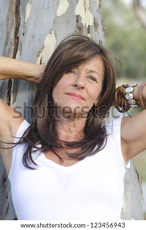 Portrait of beautiful looking middle aged woman leaning relaxed with arms up and happy smile against tree in park, with blurred background.