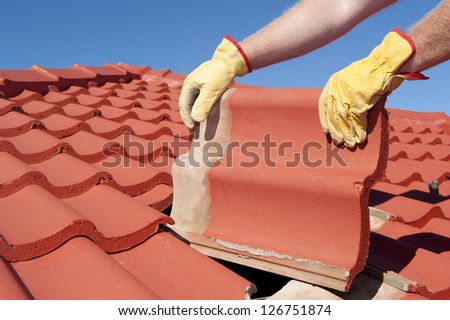 Roof repairs, worker with yellow gloves replacing red tiles or shingles on house with blue sky as background and copy space.