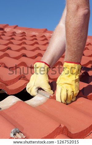 Roof repair, worker with yellow gloves replacing red tiles or shingles on house with blue sky as background and copy space.