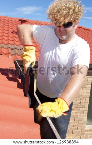 Handyman, worker cleaning gutter on house with shovel, roof with red tiles and shingles and blue sky as background.