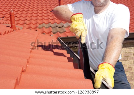 Handyman, worker cleaning gutter on house with shovel, roof with red tiles and shingles  as background.