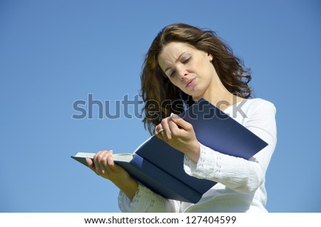 Young beautiful student girl thinking with finger at lips and with a big blue science book on the head isolated in front of blue sky with copy space